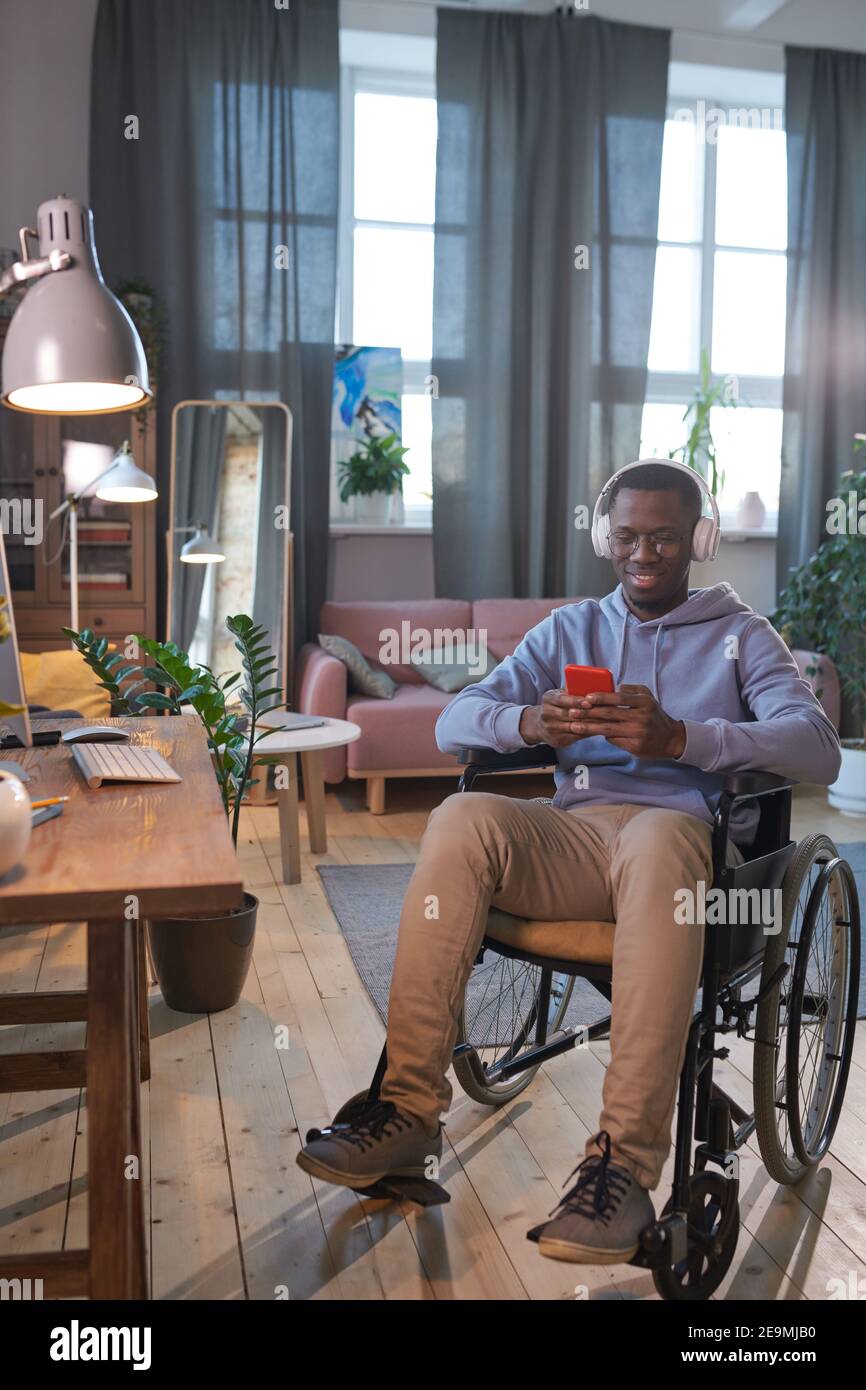 Uomo africano disabile seduto su sedia a rotelle e utilizzando il suo cellulare telefono mentre si è seduti a casa Foto Stock