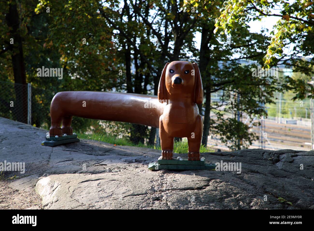 Lunga panca marrone a forma di dachshund situata in un parco per cani a Helsinki, Finlandia. Fotografato Sep, 2019. La panchina a forma di cane è su una roccia fronte di alberi Foto Stock