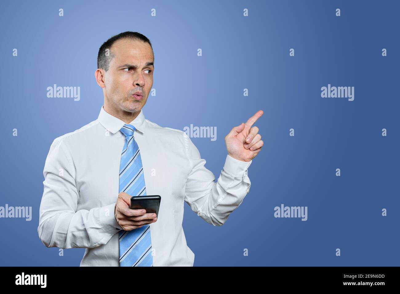 Uomo brasiliano maturo, in abiti formali, puntando il dito verso sinistra e facendo un volto. Sfondo blu. Foto Stock