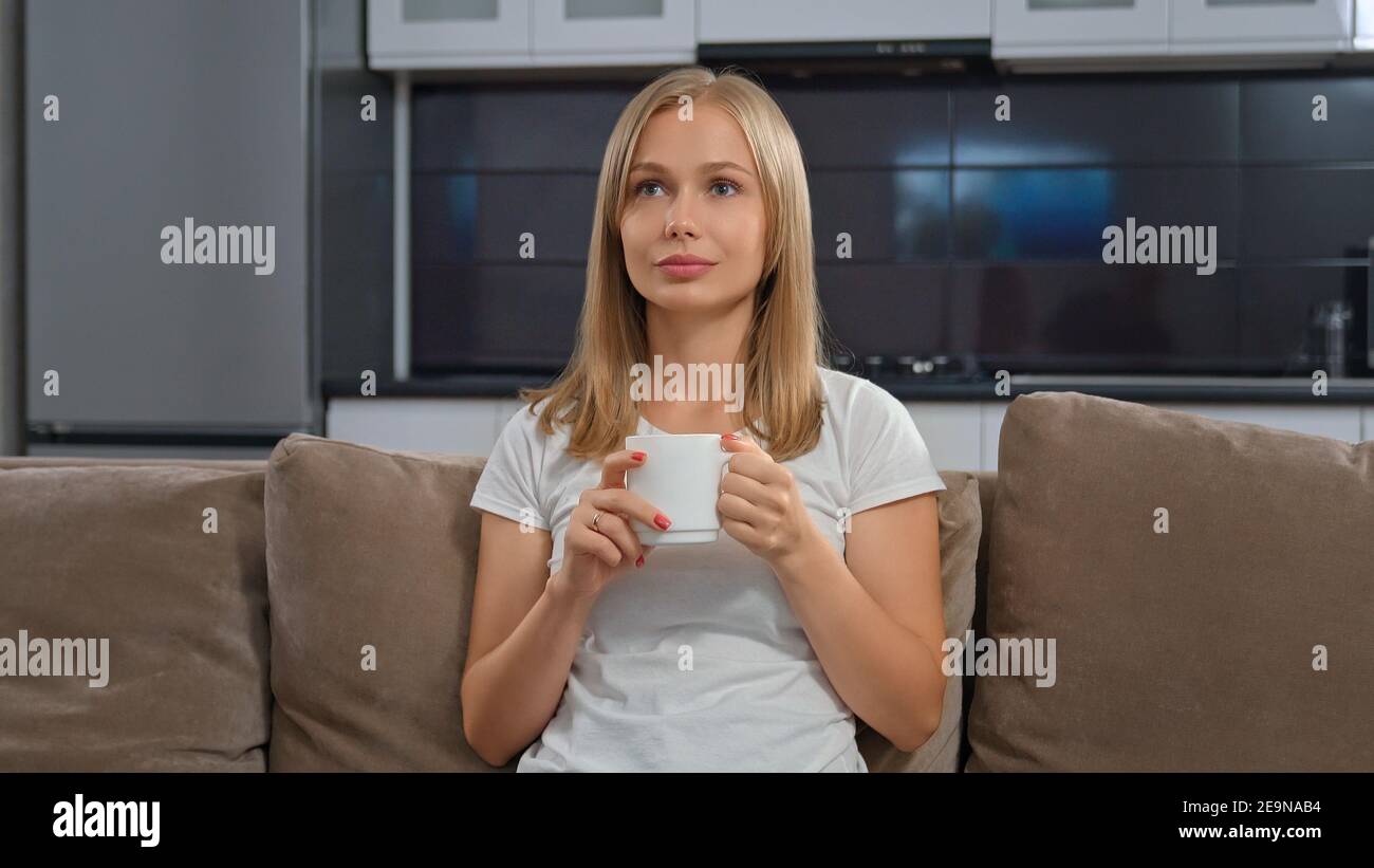 Bella donna seduta sul divano e guardando la TV. Biondo che tiene la tazza in mani e sorridente. Concetto tranquillo fine settimana a casa. Foto Stock