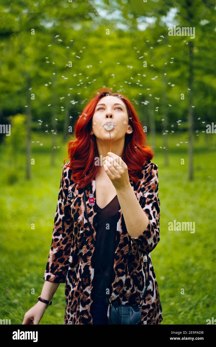 Giovane donna dai capelli rossi soffia via un dente di leone. Bella ragazza in un elegante giacca stampa leopardo. Estate atmosfera romantica. Passeggiata nel Parco. Sfondo verde sfocato. Godetevi la natura. Foto Stock