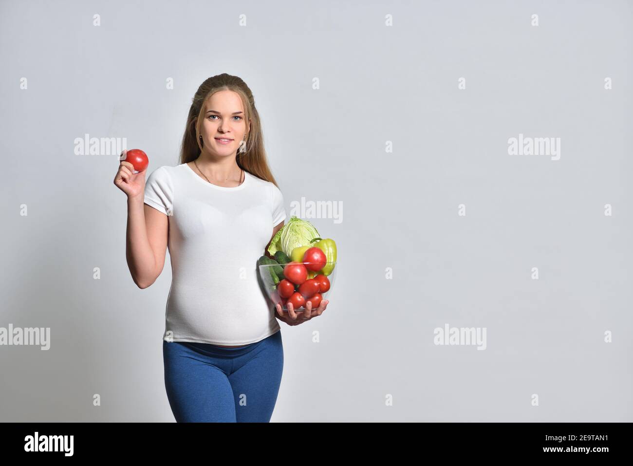 Donna bionda incinta che tiene verdure fresche in una ciotola e. pomodoro in mano Foto Stock