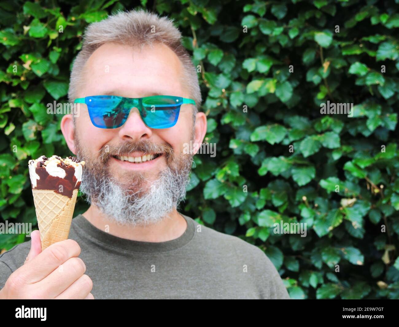 Un bell'uomo bearded in occhiali da sole tiene un gelato cono in mano e sorride Foto Stock