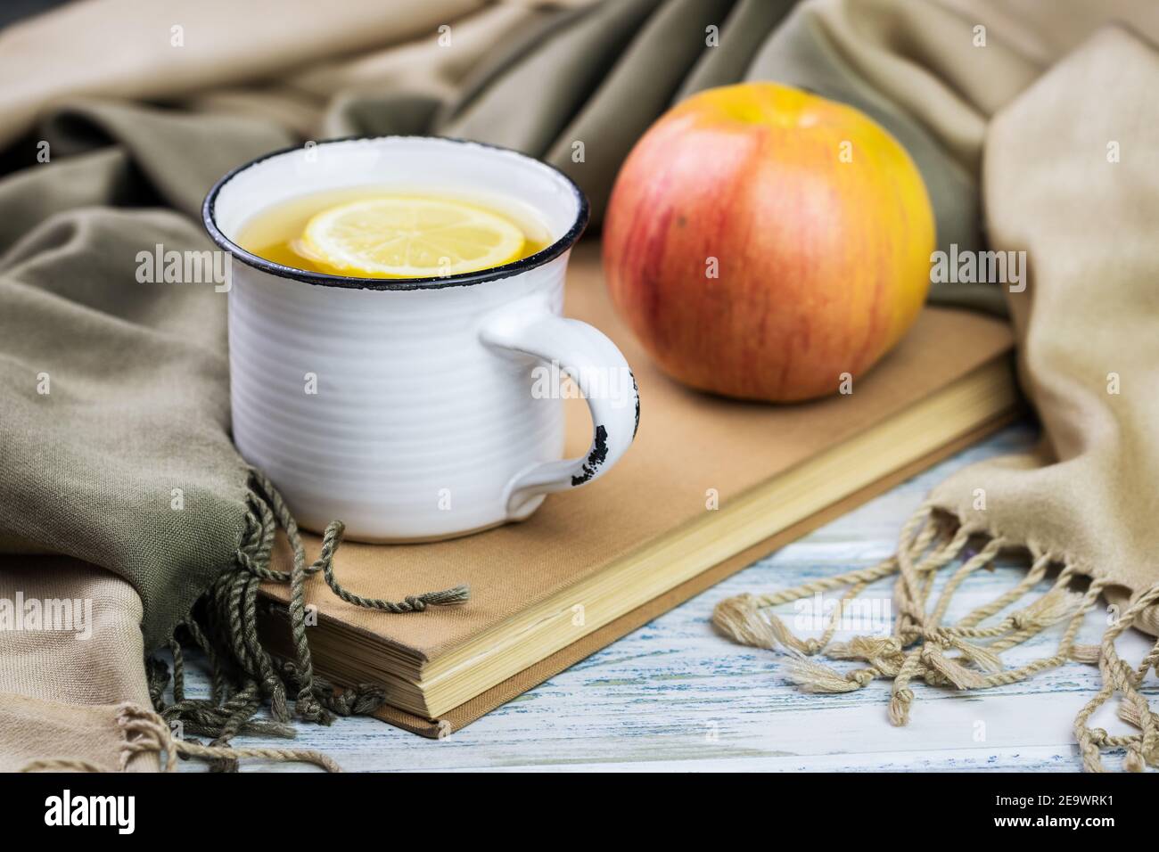 Tazza da tè e mela su libro coprendo da sciarpa, ancora vita su tavola di legno bianco. Frutta fresca biologica e bevande calde rinfrescanti. Foto Stock