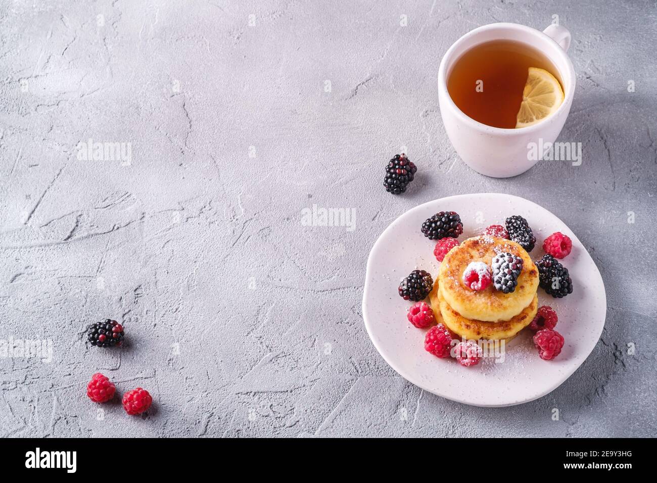 Frittelle al formaggio e zucchero in polvere, frittelle di cagliata dessert con lampone e frutti di bosco in piatto vicino alla tazza di tè caldo con fetta di limone Foto Stock