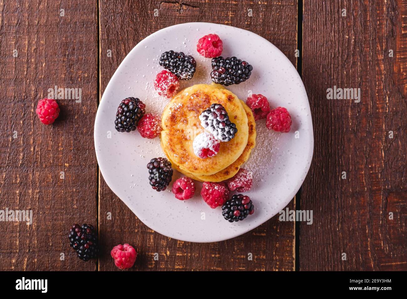 Frittelle di formaggio e zucchero in polvere, frittelle di cagliata dessert con lampone e frutti di bosco in piatto su sfondo di legno marrone scuro, top Foto Stock