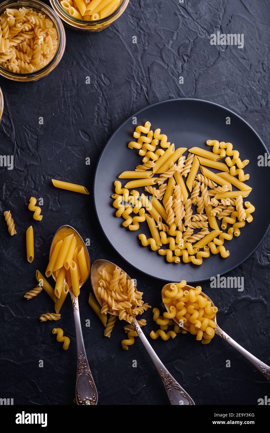 Tre cucchiai di posate, vasetti di vetro e piatto con una varietà di pasta di grano dorato cruda su sfondo nero scuro testurizzato, vista dall'alto Foto Stock