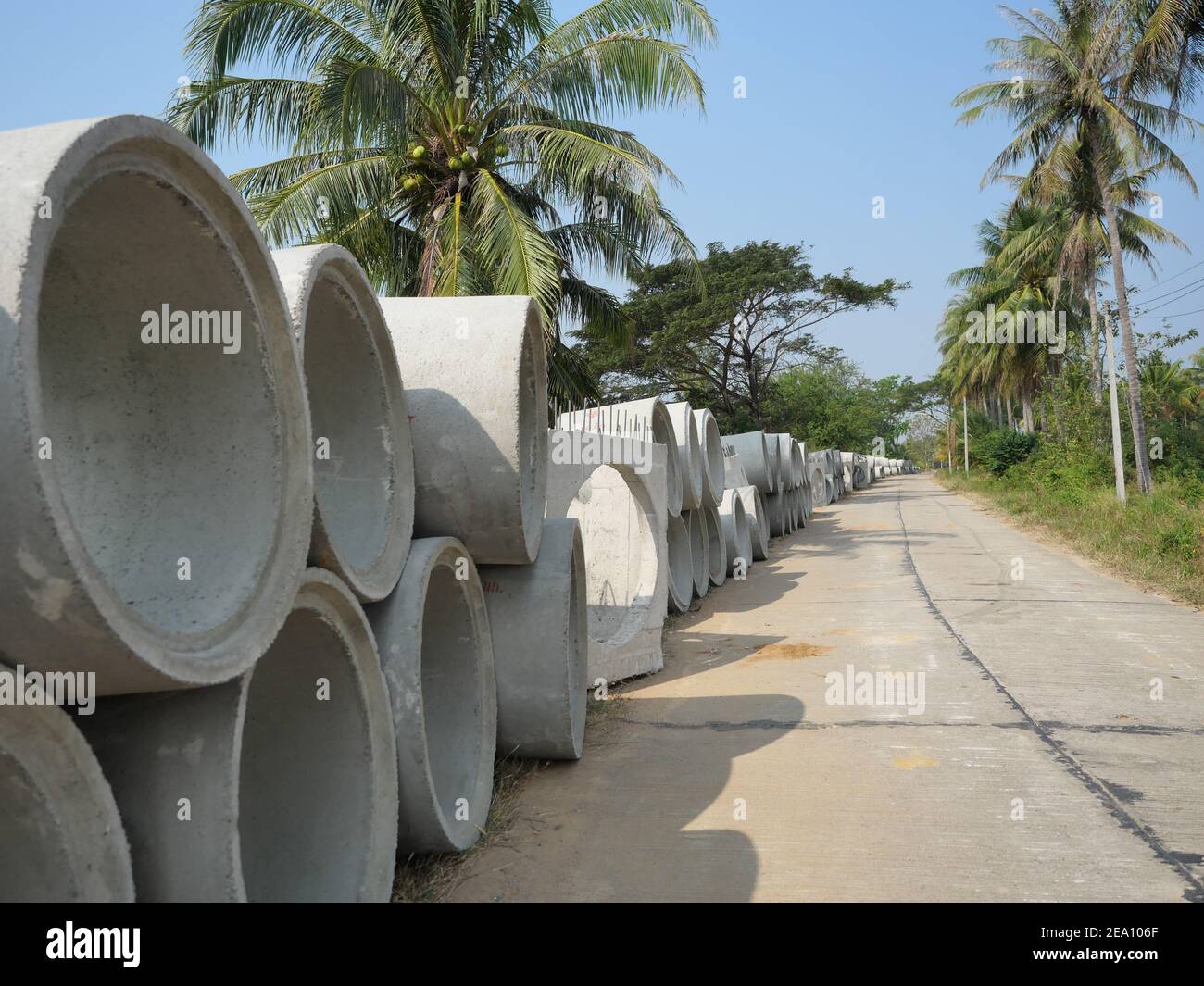 Tubi fognari in cemento sono accatastati in cantiere a bordo strada in Thailandia, strada con palme da cocco e cielo blu sullo sfondo Foto Stock