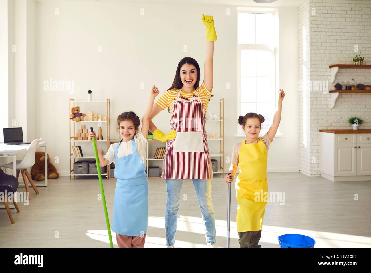 Mamma con due figlie che puliscono la casa in piedi con le braccia alzate fingendo di essere supereroi. Foto Stock
