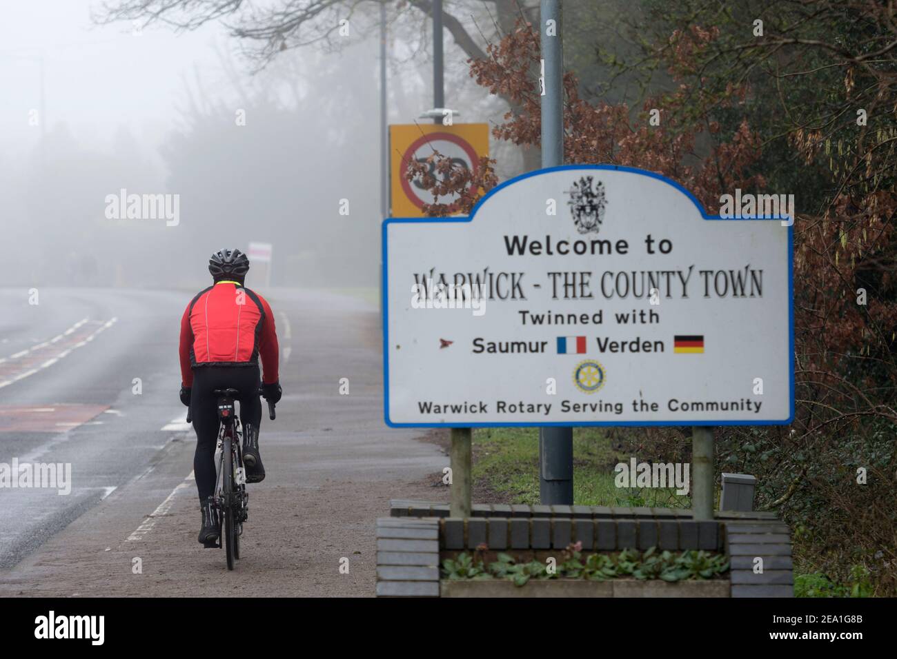 Un ciclista in un giorno foggy in inverno che passa un cartello di benvenuto a Warwick, Warwickshire, Inghilterra, Regno Unito Foto Stock