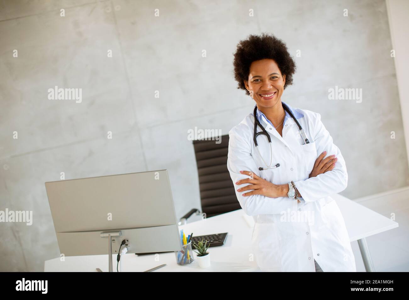 Donna Africana americana medico indossando camice bianco con stetoscopio in piedi dalla scrivania in ufficio Foto Stock