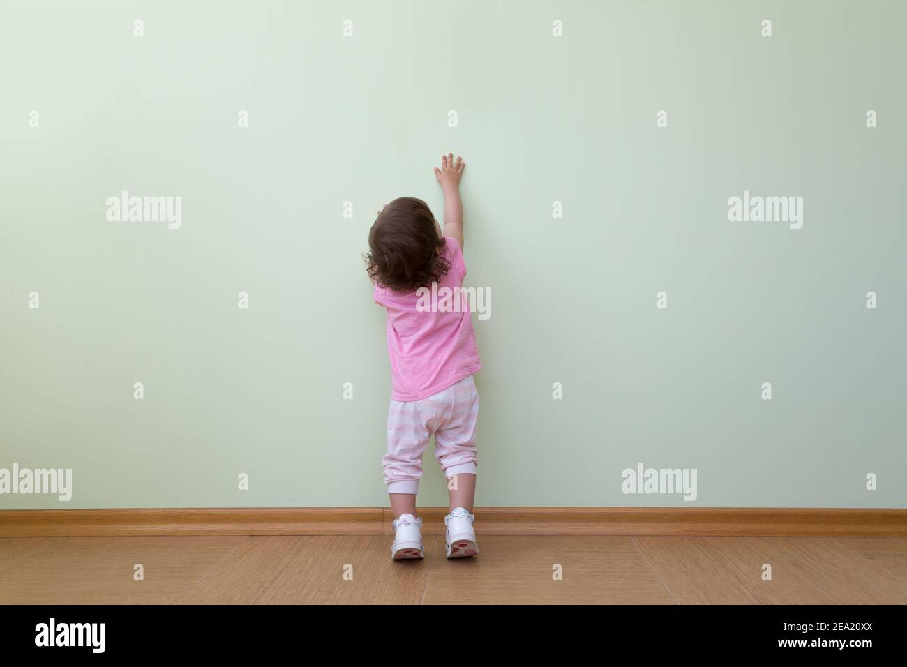 Adorabile bambina in una blusa rosa e pantaloni a righe si trova di fronte a una parete verde e guarda in su. Il concetto di scoperta, il desiderio di conoscenza Foto Stock