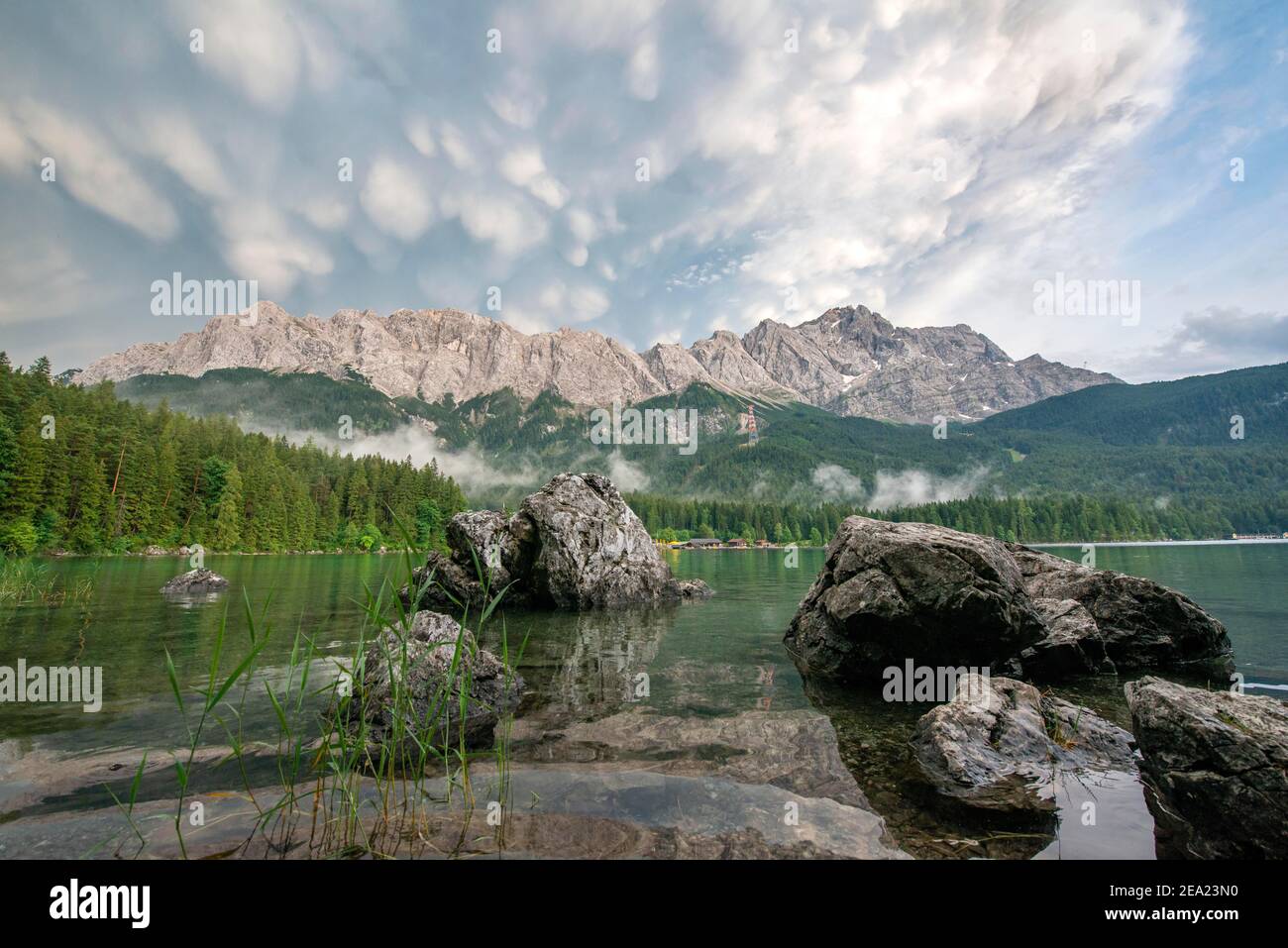 Rocce sulla riva, lago Eibsee di fronte al massiccio Zugspitze con Zugspitze, drammatiche nubi mammaten, Wetterstein, vicino Grainau, Upper Foto Stock