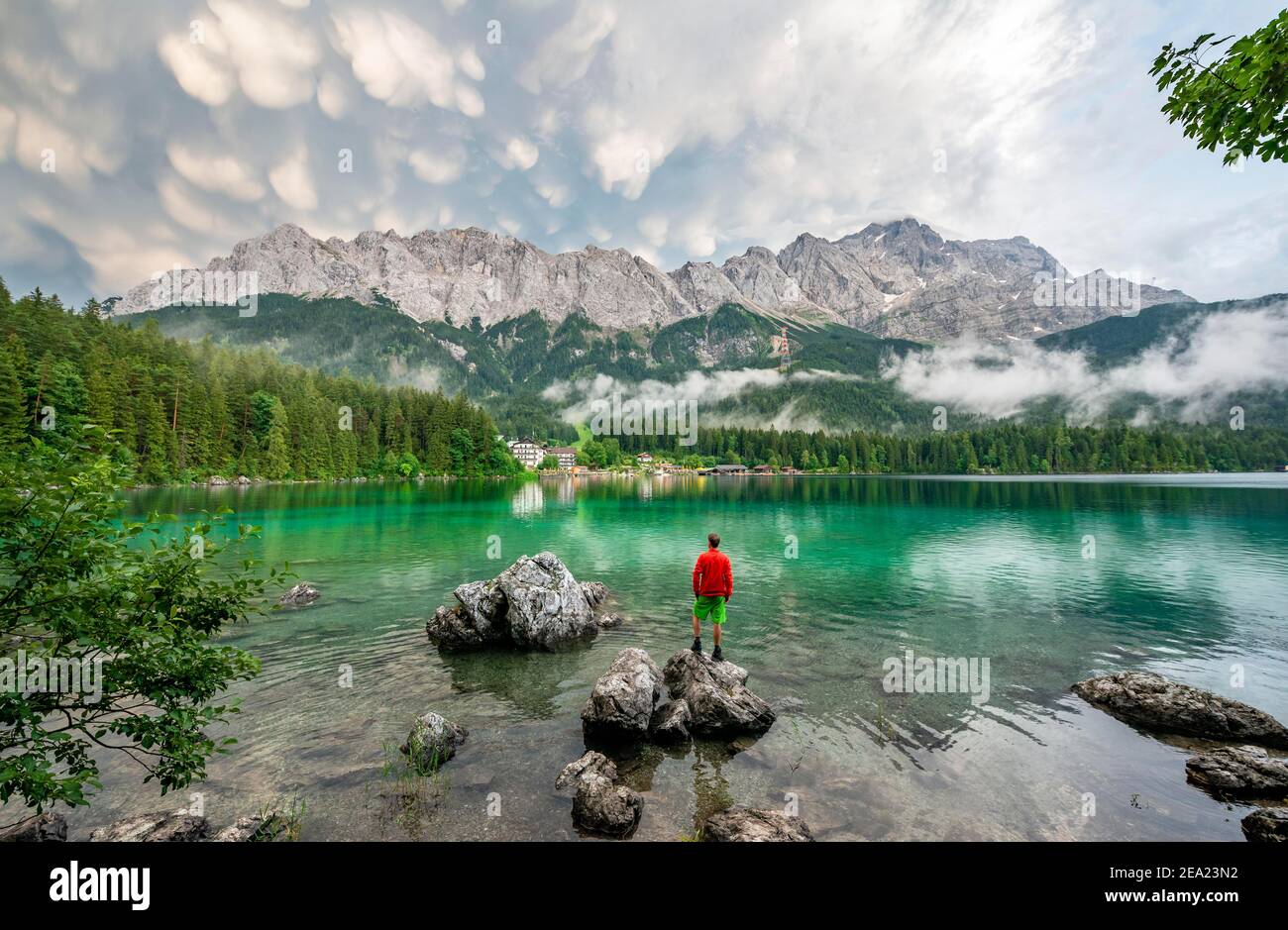 Giovane uomo in piedi su una roccia sulla riva, lago Eibsee di fronte al massiccio Zugspitze con Zugspitze, drammatiche nuvole mammaten, Wetterstein gamma, vicino Foto Stock