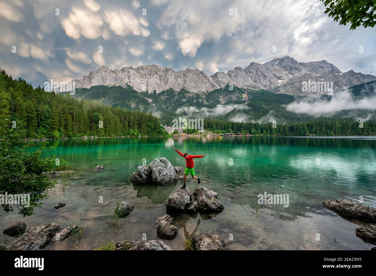 Il giovane salta da roccia a roccia, il lago Eibsee di fronte al massiccio di Zugspitze con Zugspitze, drammatiche nuvole mammaten, Wetterstein, vicino Foto Stock