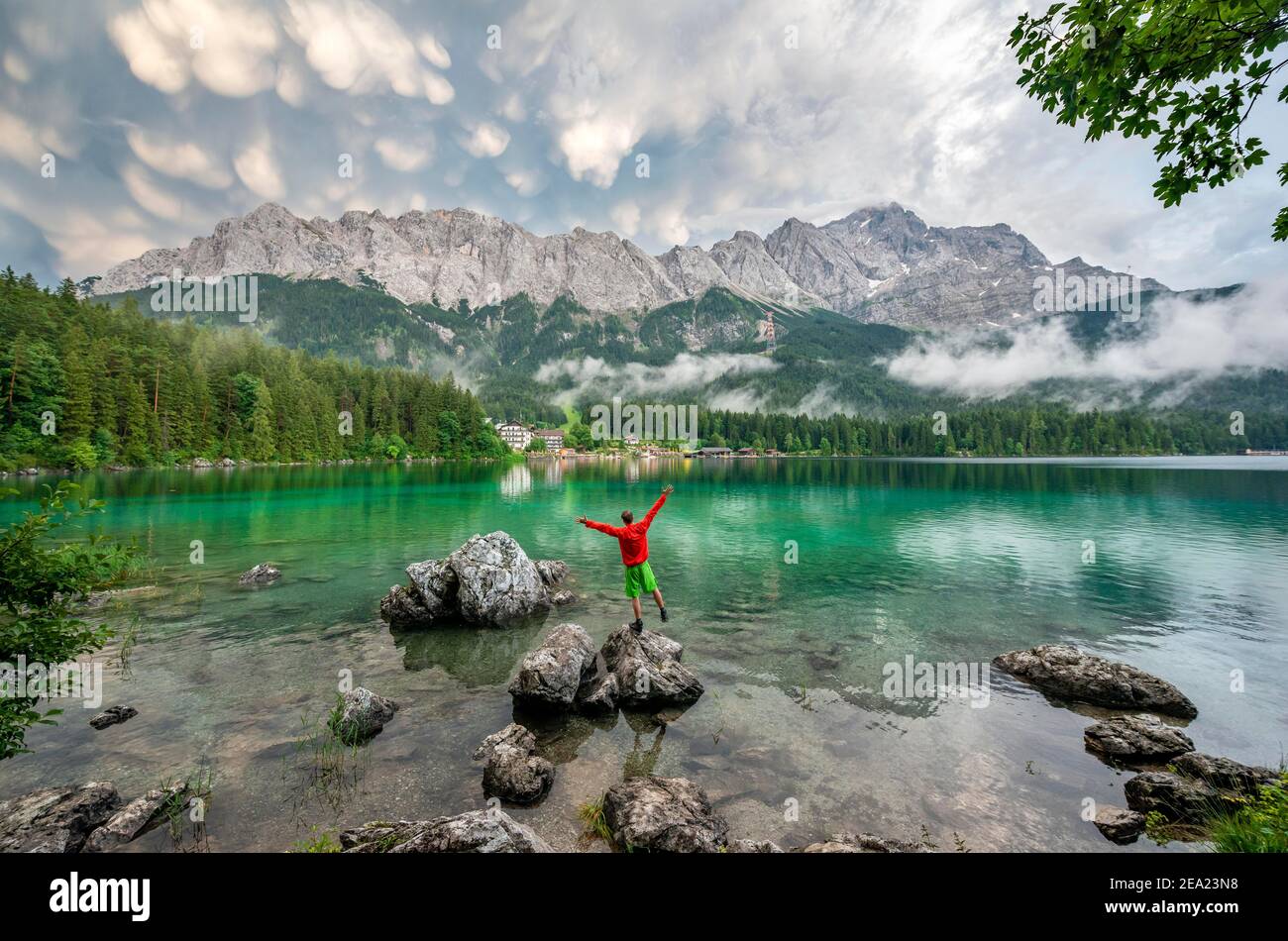 Il giovane attacca le braccia in aria e si erge su una roccia sulla riva, il lago Eibsee di fronte al massiccio di Zugspitze con Zugspitze, drammatico Mammaten Foto Stock