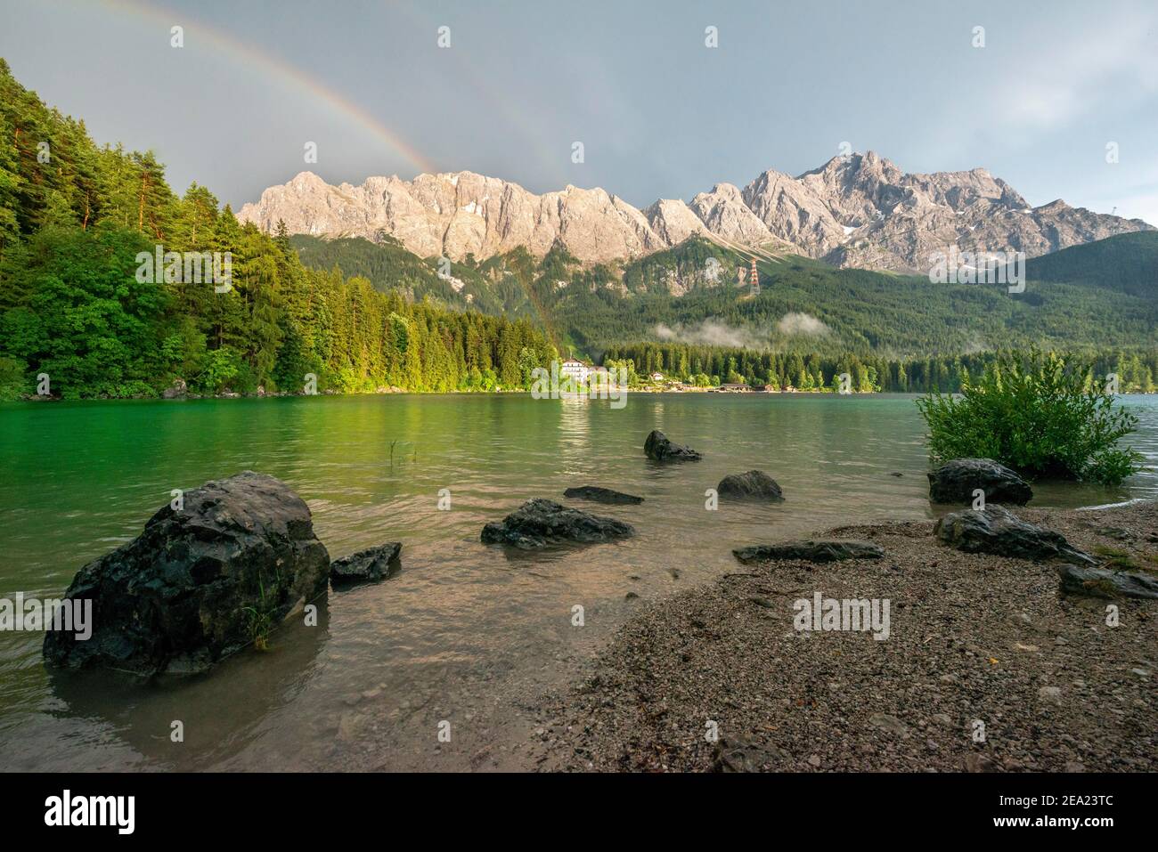 Rocce sulla riva, lago di Eibsee di fronte al massiccio di Zugspitze con Zugspitze con arcobaleno, dietro tuonuvole, catena montuosa di Wetterstein, vicino Grainau Foto Stock