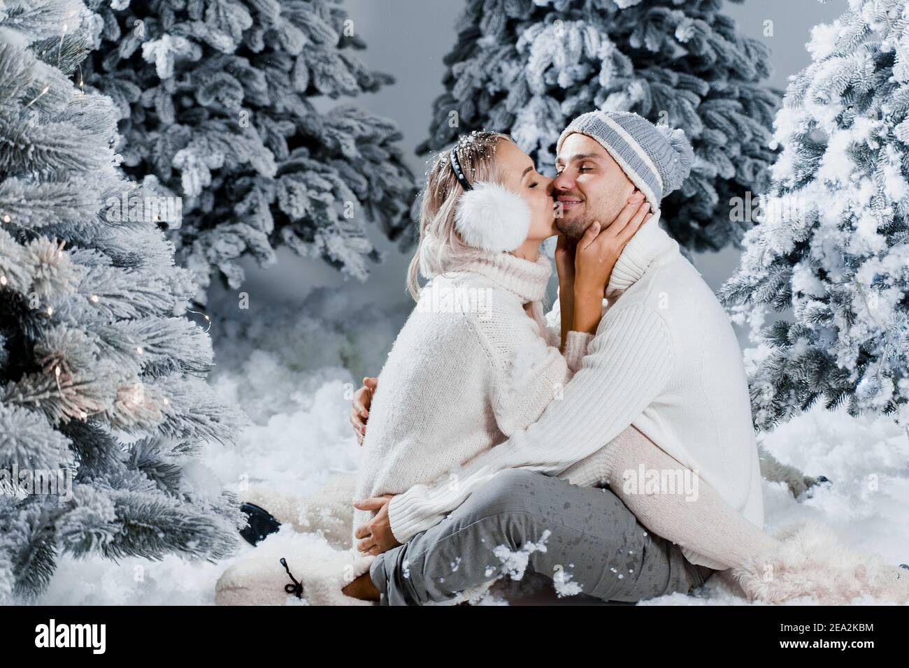 Storia d'amore invernale con due cuffie in pelliccia, cappelli, pullover  bianchi. Felice giovane coppia abbracci e bacio vicino alberi di natale  alla vigilia di nuovo yea Foto stock - Alamy