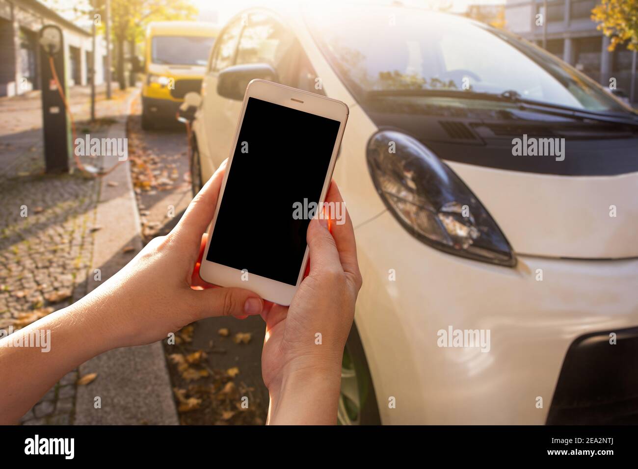 Mani con lo smartphone sullo sfondo di un'auto a noleggio presso la stazione di ricarica per i veicoli elettrici. Condivisione dell'auto. Foto Stock