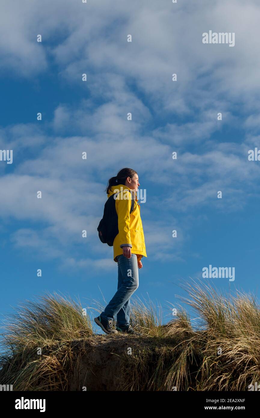 Donna escursionista che cammina lungo alte dune di sabbia. Giacca gialla e zaino contro un cielo blu. Foto Stock