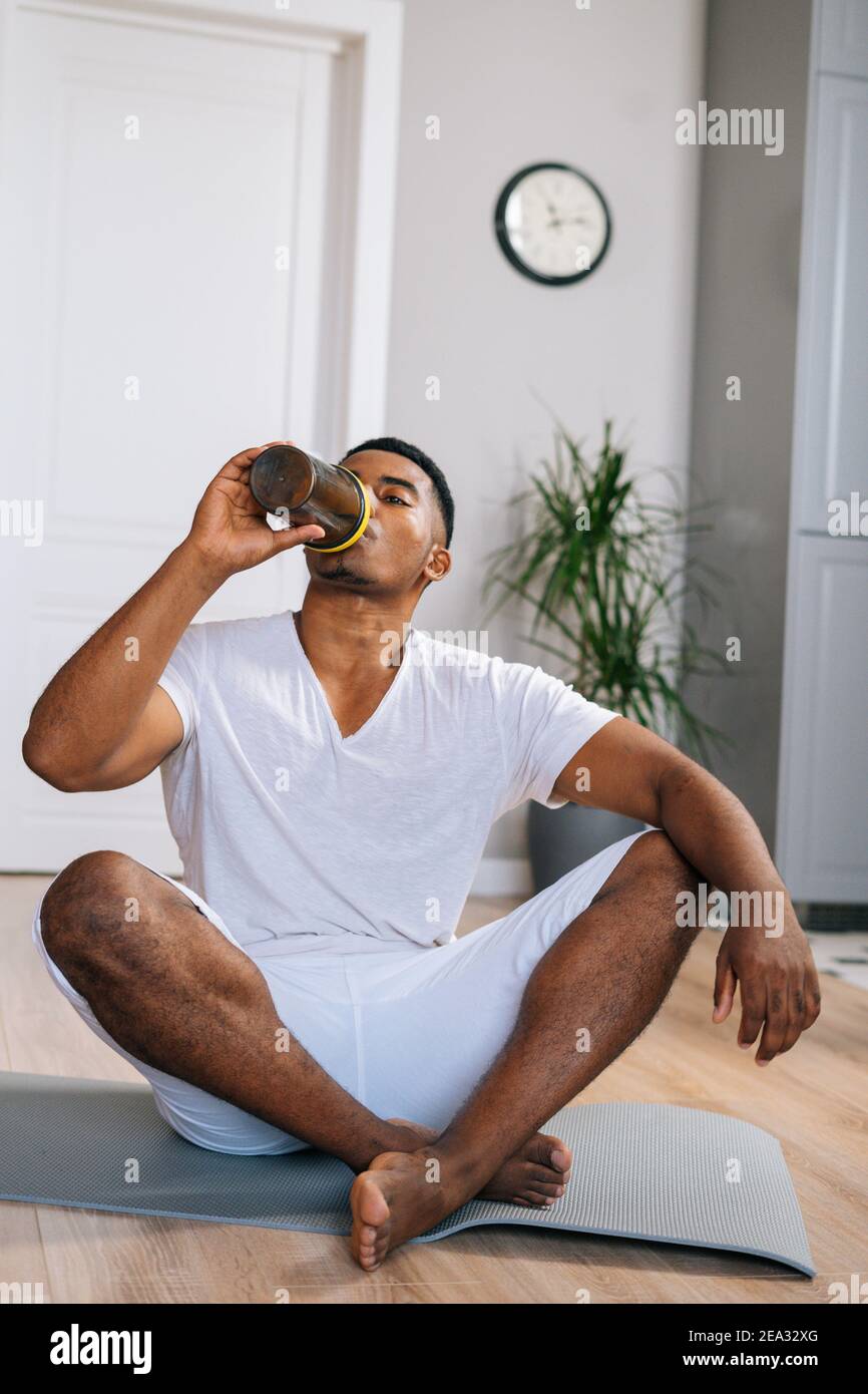 L'uomo afroamericano si rilassa sul tappeto da ginnastica dopo l'allenamento e beve una bottiglia di acqua dolce. Foto Stock