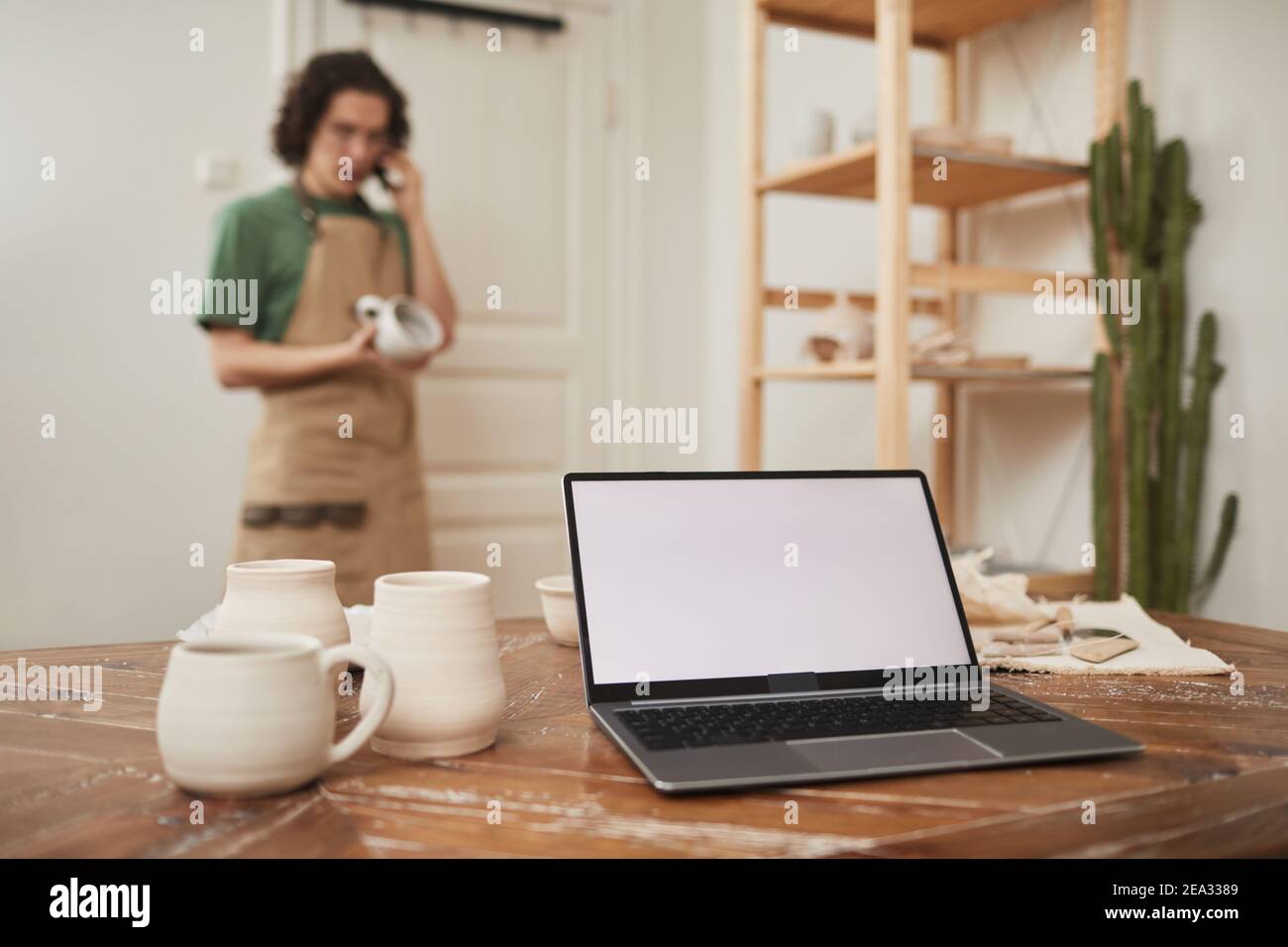 Ancora vita di computer portatile con schermo bianco vuoto sul tavolo in officina, con blured artigiano maschile parlare per telefono in background, piccola impresa e entre Foto Stock