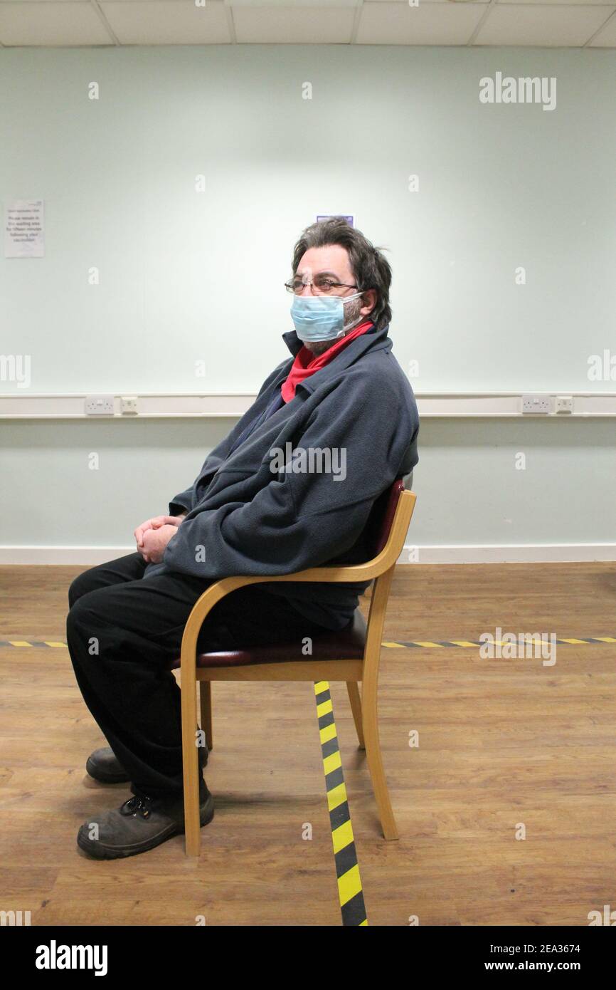 Un uomo maturo indossando occhiali e una maschera sedette dentro un ospedale sala d'attesa dopo aver ricevuto la sua vaccinazione coviale Foto Stock