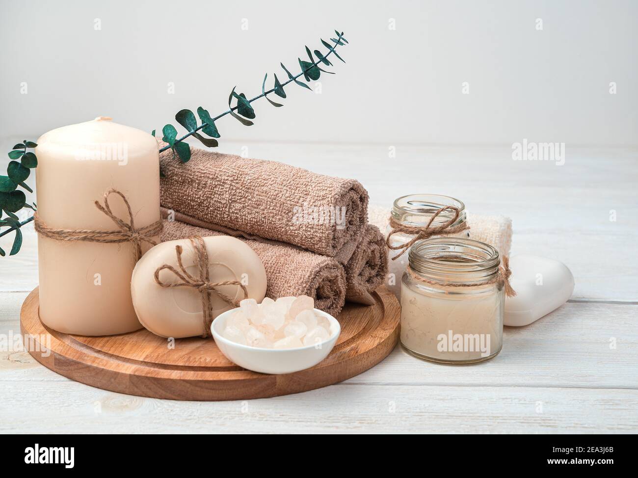 Candele, sapone e un set di asciugamani marroni con un ramo di eucalipto su uno sfondo di legno bianco. Foto Stock