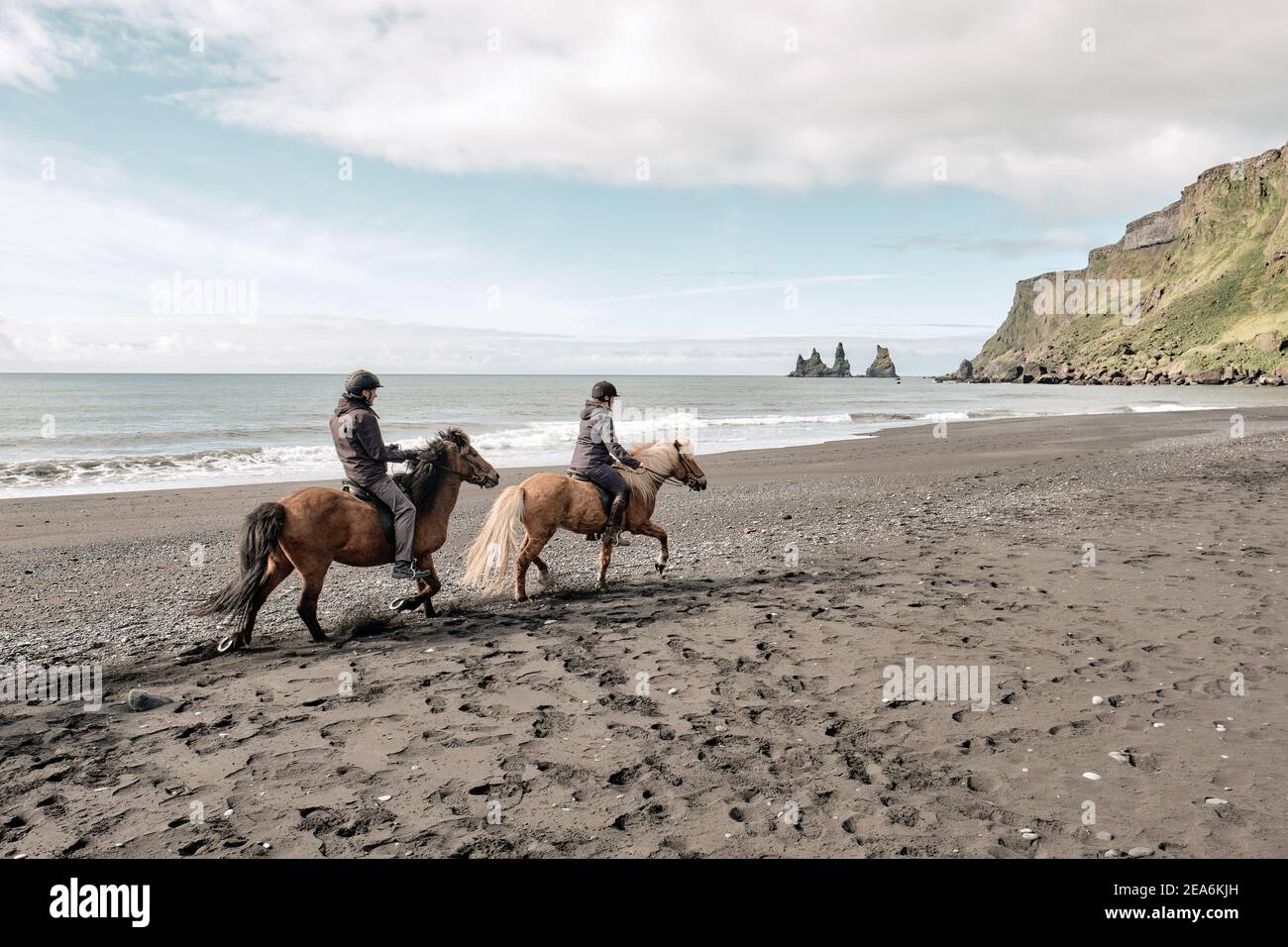 Turisti in un giro a cavallo islandese sulla sabbia nera di Reynisdrangar spiaggia con Reynisdrangar mare stack dietro - Islanda equitazione vacanza di attività Foto Stock