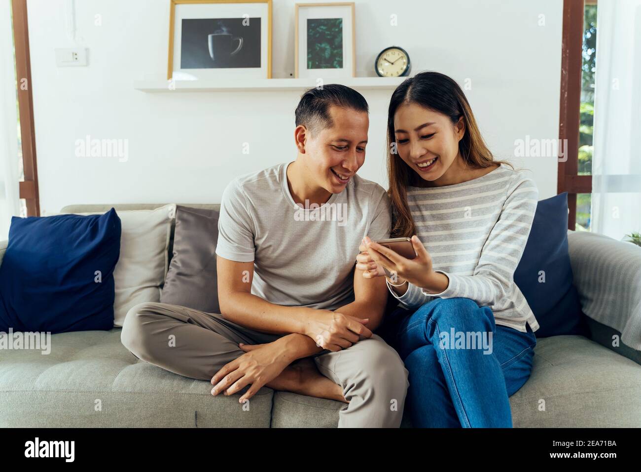 Anni 30 giovane adulto asiatico uomo e donna giocare un telefono cellulare insieme si siede sul divano in un accogliente soggiorno a casa. Felice utente di Internet di coppia in abiti casual sul divano. Concetto di utilizzo della tecnologia in Asia Foto Stock