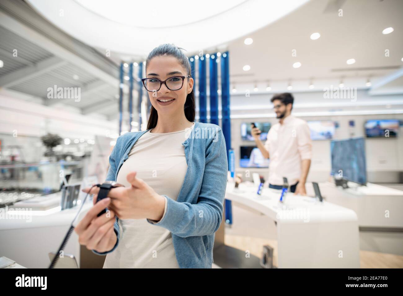 Donna che tiene in mano un orologio intelligente mentre si trova in un negozio di tecnologia e si guarda in macchina fotografica. Foto Stock