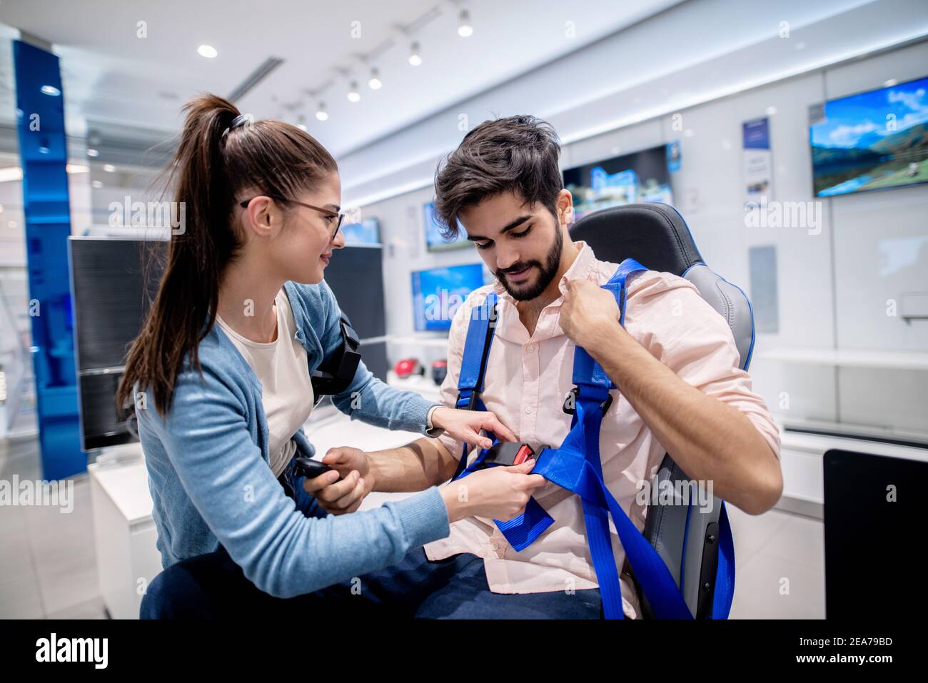 Donna che aiuta l'uomo a allacciare la cintura di sicurezza per il sedile VR. Negli schermi al plasma a sfondo. Foto Stock