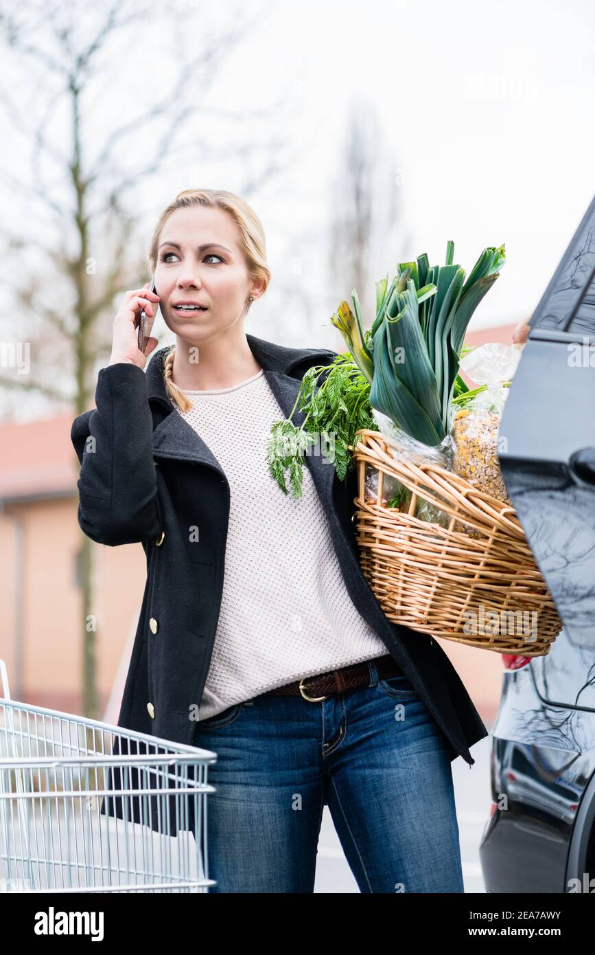 Donna che usa il telefono mentre immagazzina i generi alimentari nel bagagliaio dell'automobile Foto Stock