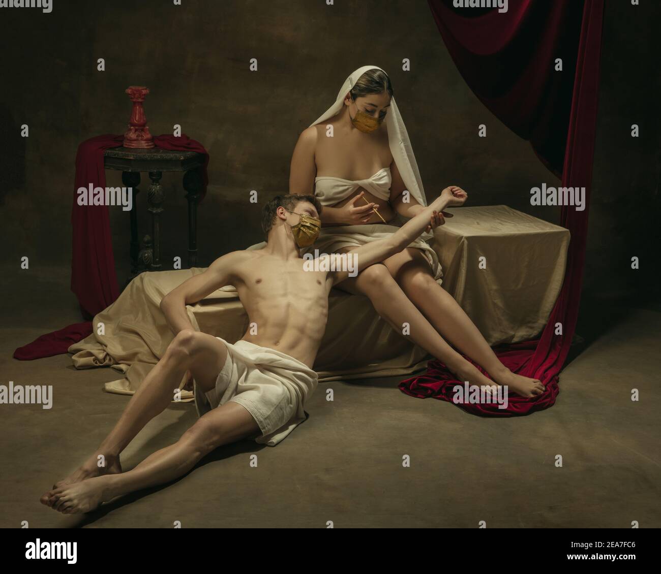 La vaccinazione sta iniziando qui. Moderno remake di opere d'arte classiche con tema coronavirus - giovane coppia medievale su sfondo scuro, di colore dorato. Concetto di coronavirus, pandemia, creatività. Foto Stock