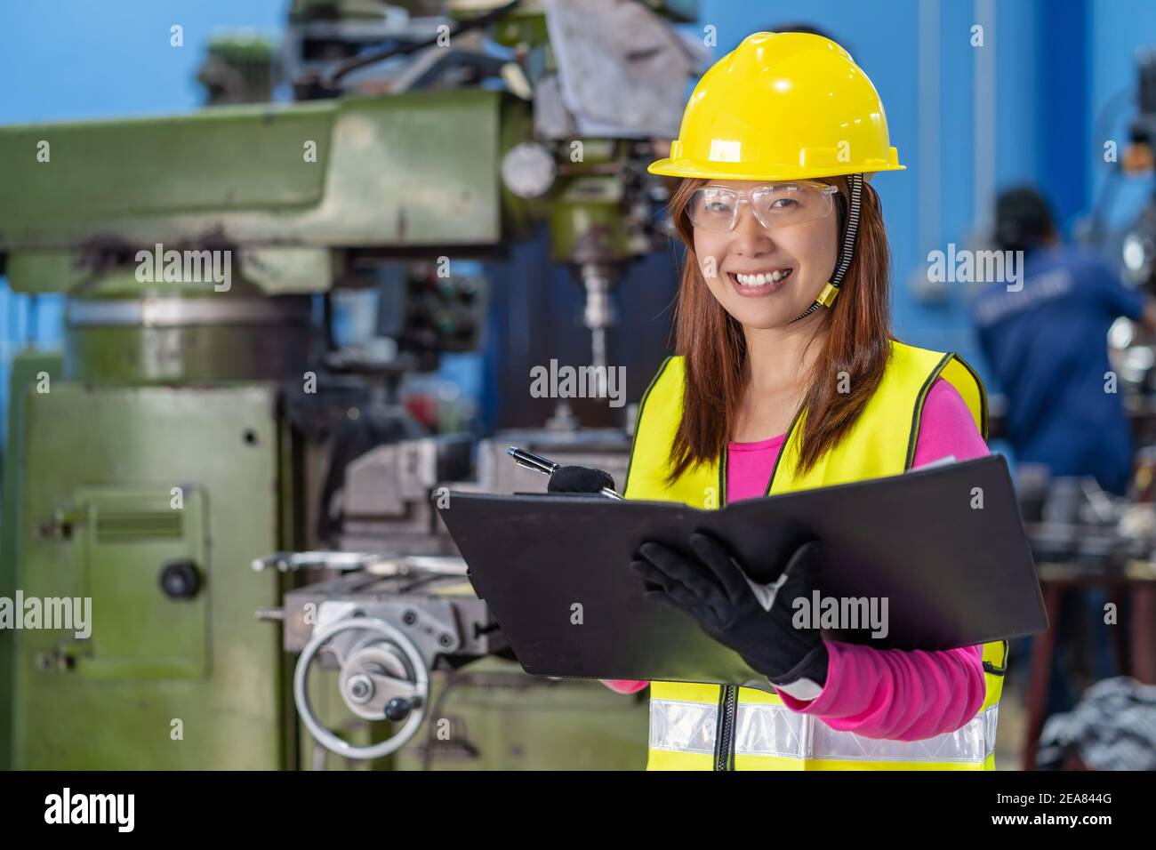 Ritratto di una donna asiatica che controlla l'elenco dei lavori in carta sulla foto sfocata del tornio e dello sfondo della fresatrice in fabbrica di metallo, Foto Stock