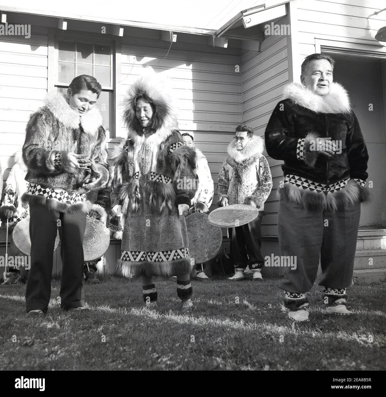 1960, storico, al confine esterno di Anchorage, Alaska, USA, al di fuori di un edificio di legno a un piano, i nativi in abito tradizionale di pelli animali e pellicce esecuzione di una danza. L'Alaska divenne il 49° stato degli Stati Uniti nel 1959, ma fu occupata da diverse popolazioni indigene per migliaia di anni prima dell'arrivo degli europei. Dato che lo Stato ha un confine marittimo con la Russia, sono stati loro a stabilirsi per primi, stabilendo ciò che è diventato noto come America russa. Le popolazioni indigene costituiscono oltre il 15% della popolazione e i nativi dell'Alaska svolgono un ruolo importante nel governo locale Foto Stock