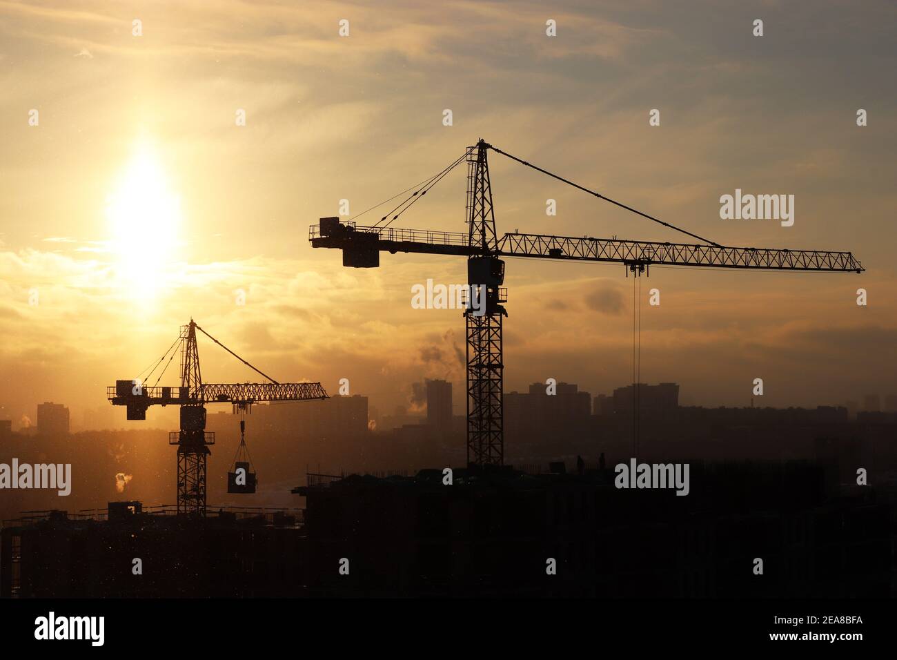 Silhouette di gru da costruzione e edificio residenziale incompiuto durante la neve sullo sfondo dell'alba. Costruzione di alloggi, condominio Foto Stock