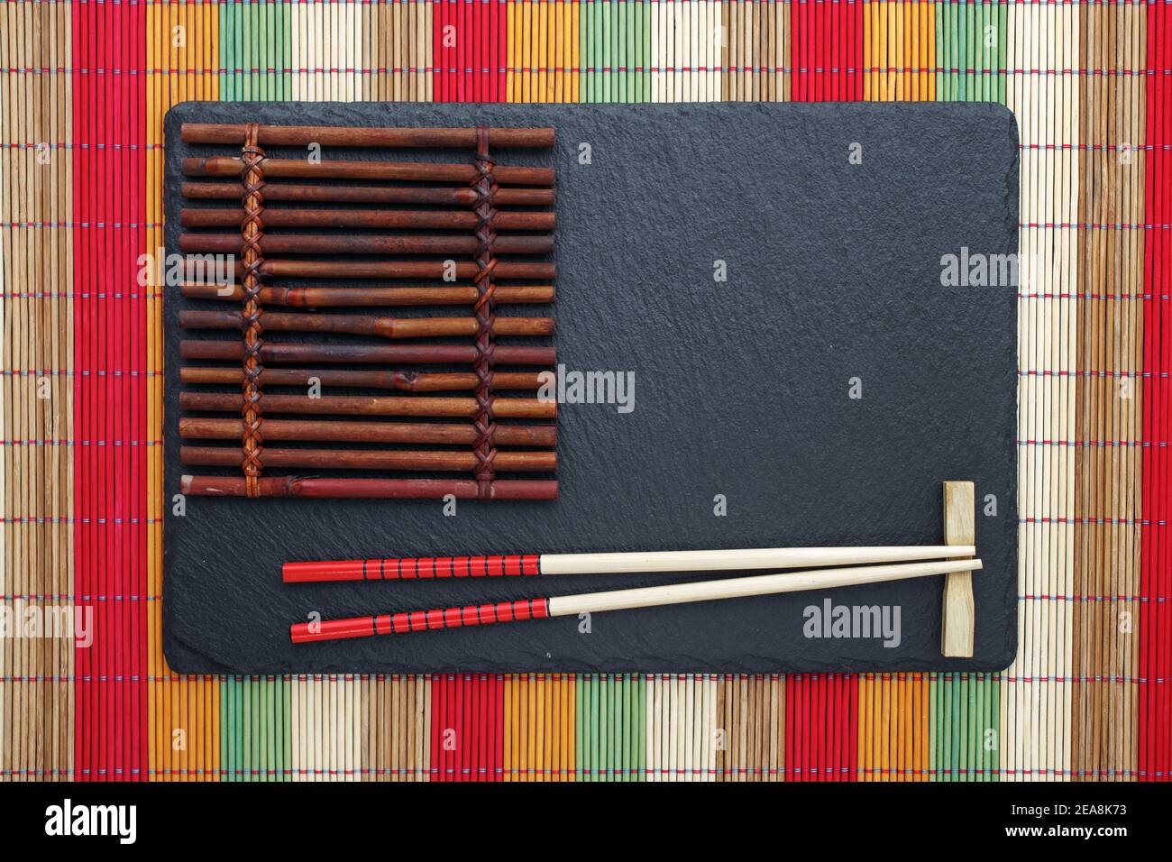 Chopsticks su piatto di ardesia nera con montagne russe su tappeto di bambù multicolore. Sfondo di cibo di stile Giapponese. Vista dall'alto con spazio per la copia Foto Stock