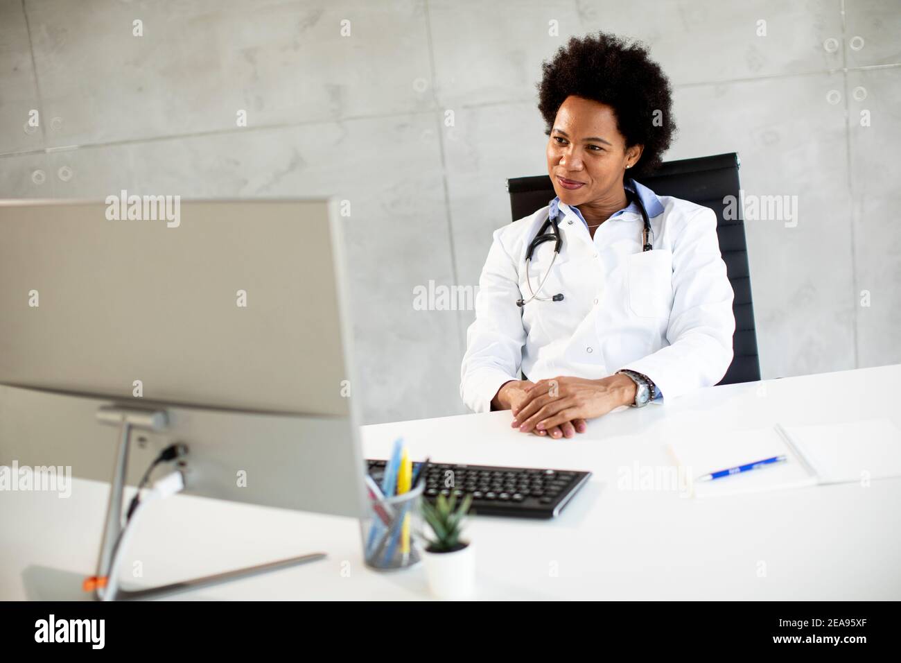 Donna Africana americana medico indossando camice bianco con stetoscopio seduta dietro la scrivania in ufficio Foto Stock