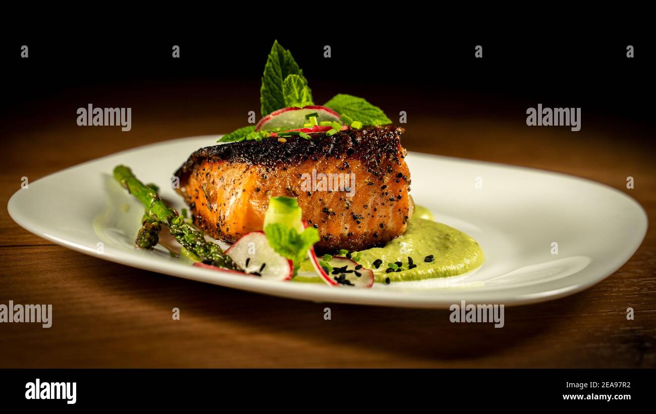 Padella croccante di salmone scottato con purea di piselli e garnishes verdi in piatto bianco. Isolato su sfondo di legno scuro Foto Stock