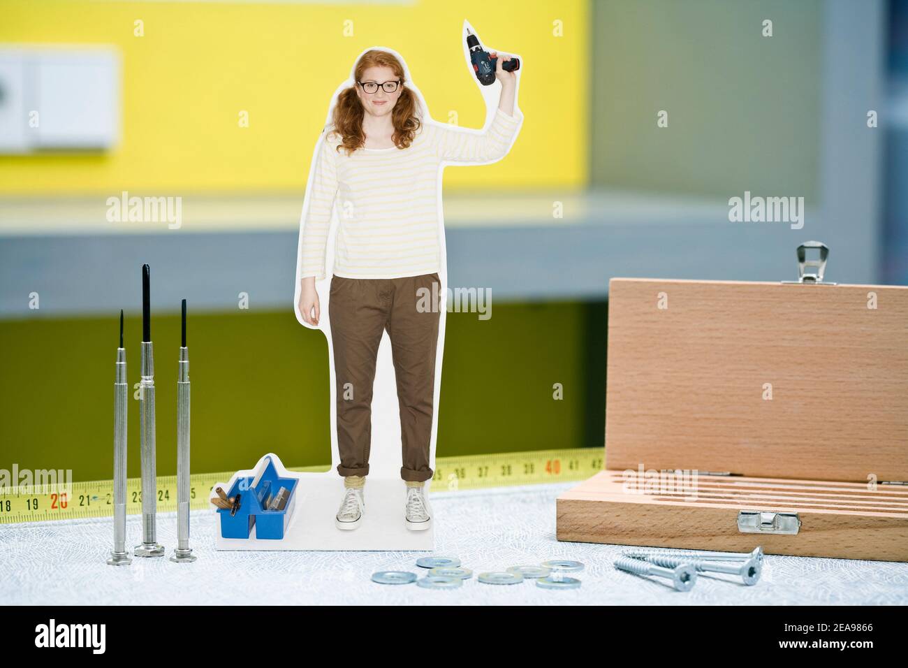 Collage fotografico, donna con trapano a batteria in mano, accanto a vari utensili, di fronte a una parete giallo-grigia, in posa vittoria Foto Stock