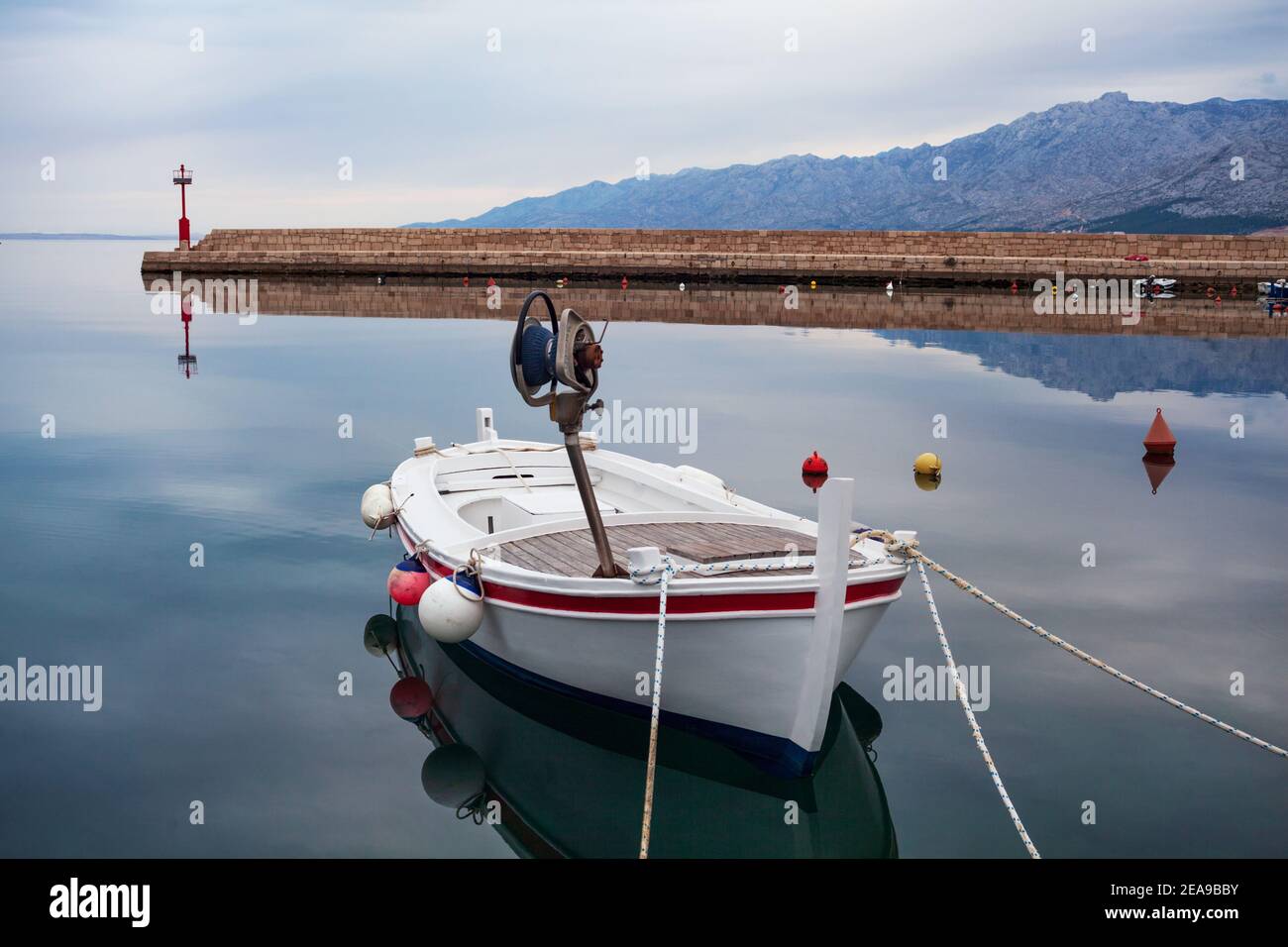 Panorama mattutino con mare calmo, barca da pesca bianca e molo con faro e montagne sullo sfondo. Foto Stock