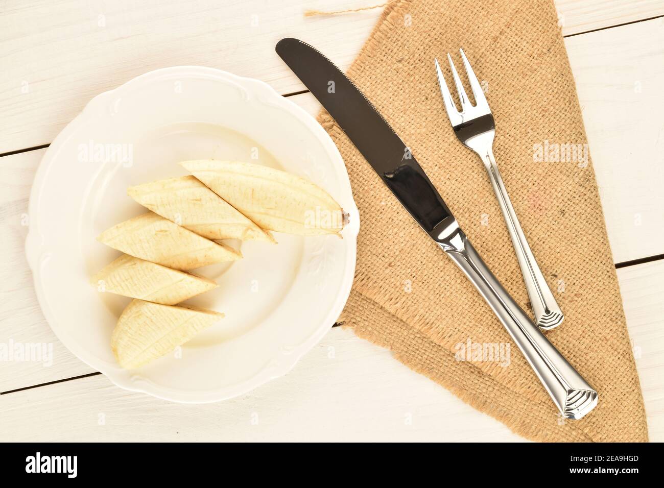 Una sbucciata e tagliata a fette, una banana matura e gustosa, su un piatto di ceramica bianca, coltelli da tavola in metallo e una forchetta su un tovagliolo di iuta, su uno sfondo Foto Stock