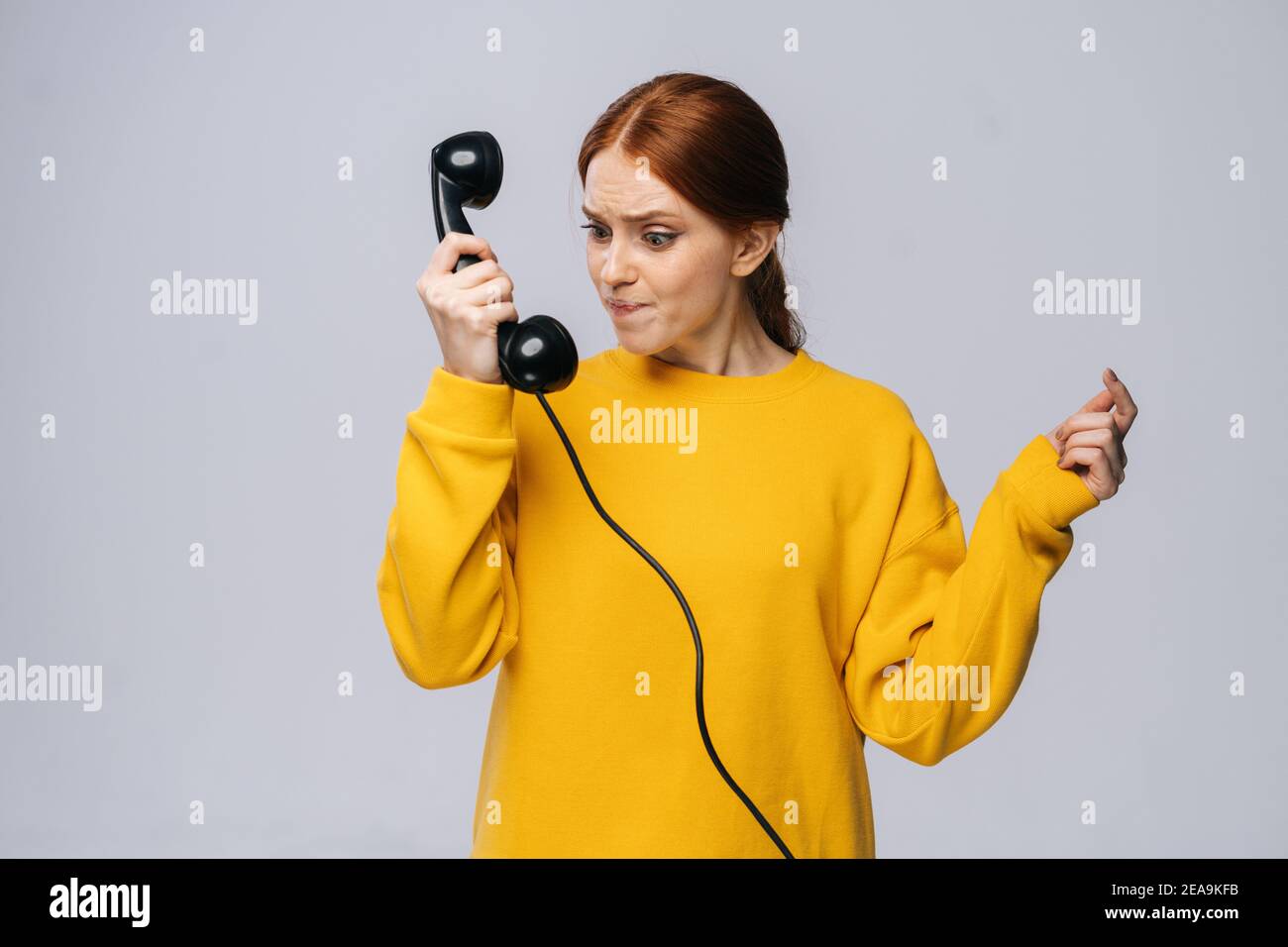 Giovane donna arrabbiata in elegante maglione che tiene ricevitore del ricevitore del ricevitore telefono retro e guardandolo Foto Stock