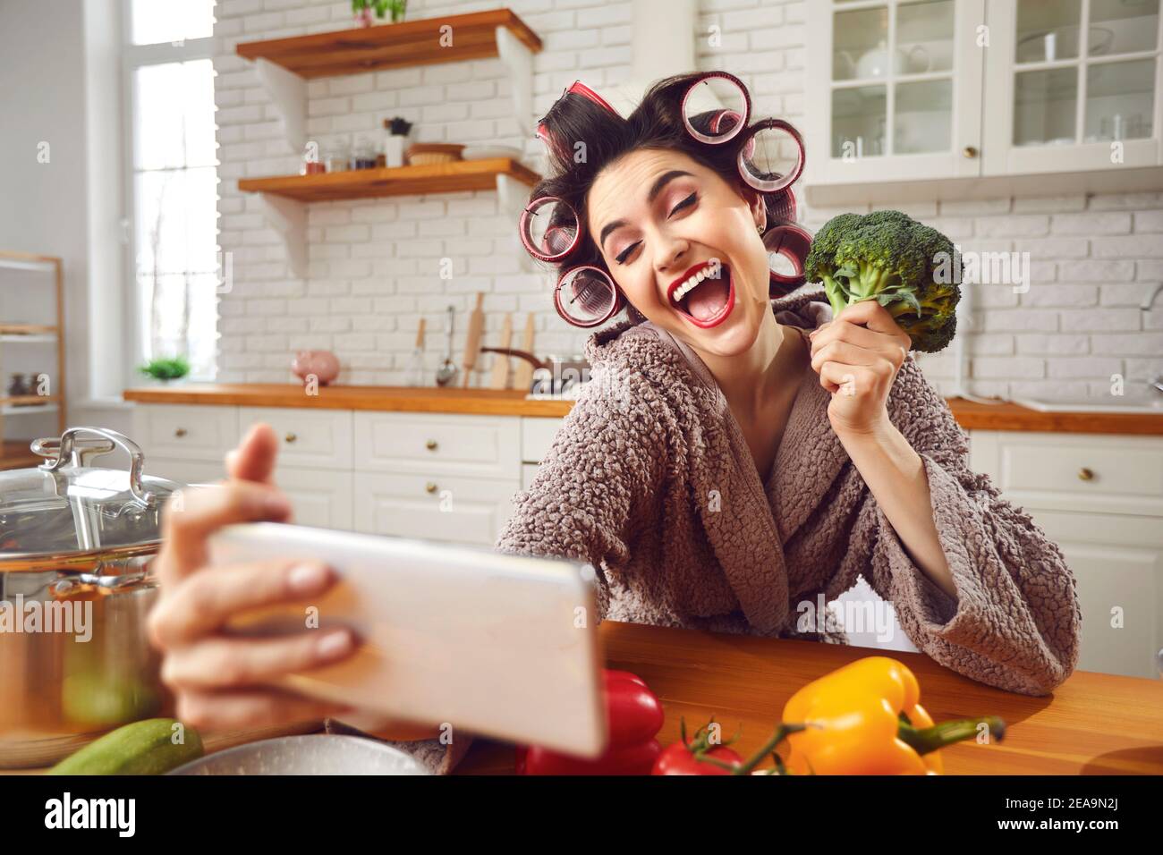 Felice giovane donna in curlers prendendo selfie divertente con broccoli testa in cucina Foto Stock