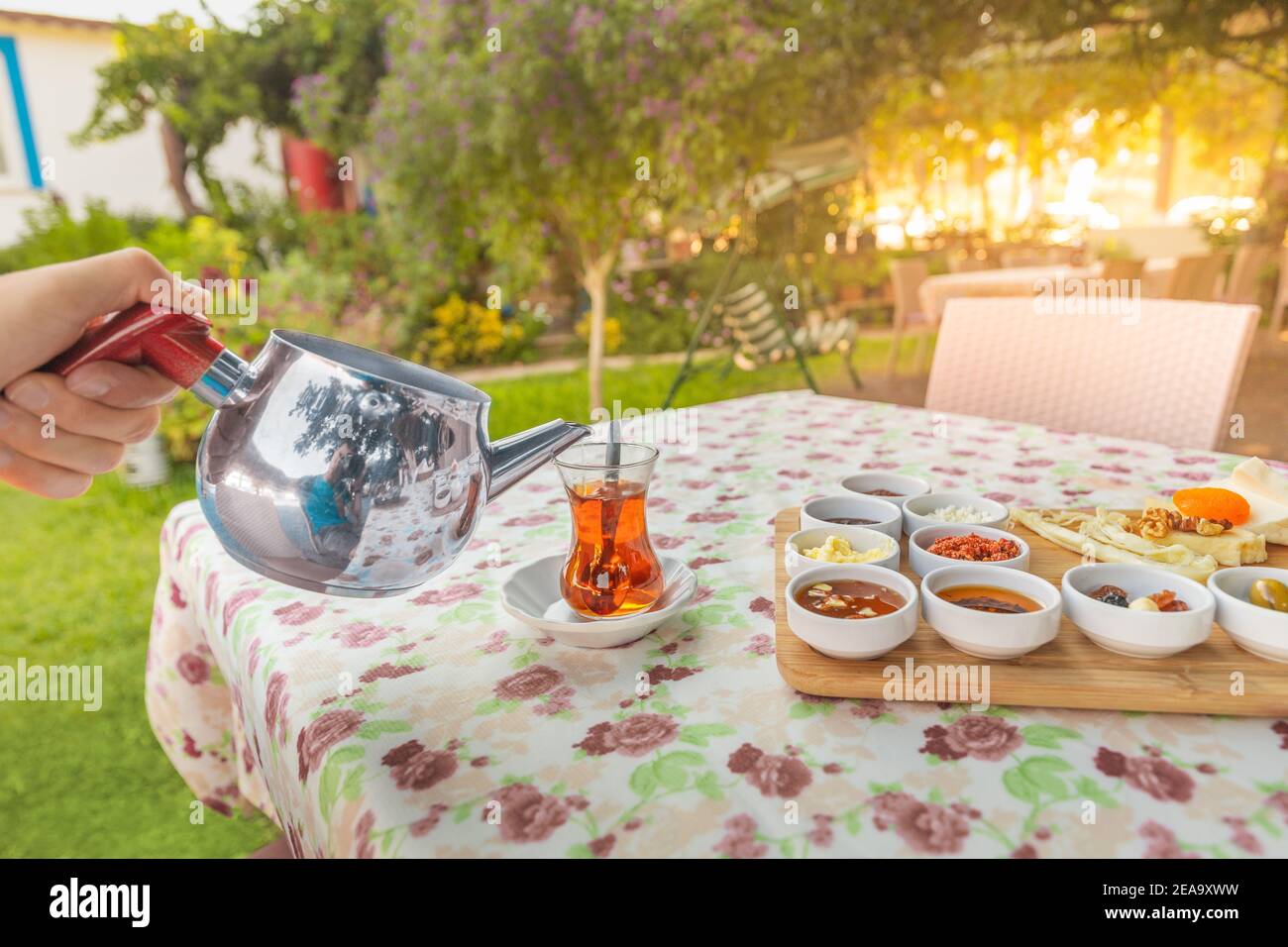 Un uomo versa il tè da una teiera in un tradizionale bicchiere turco sulla terrazza di un ristorante in un pittoresco giardino. Foto Stock