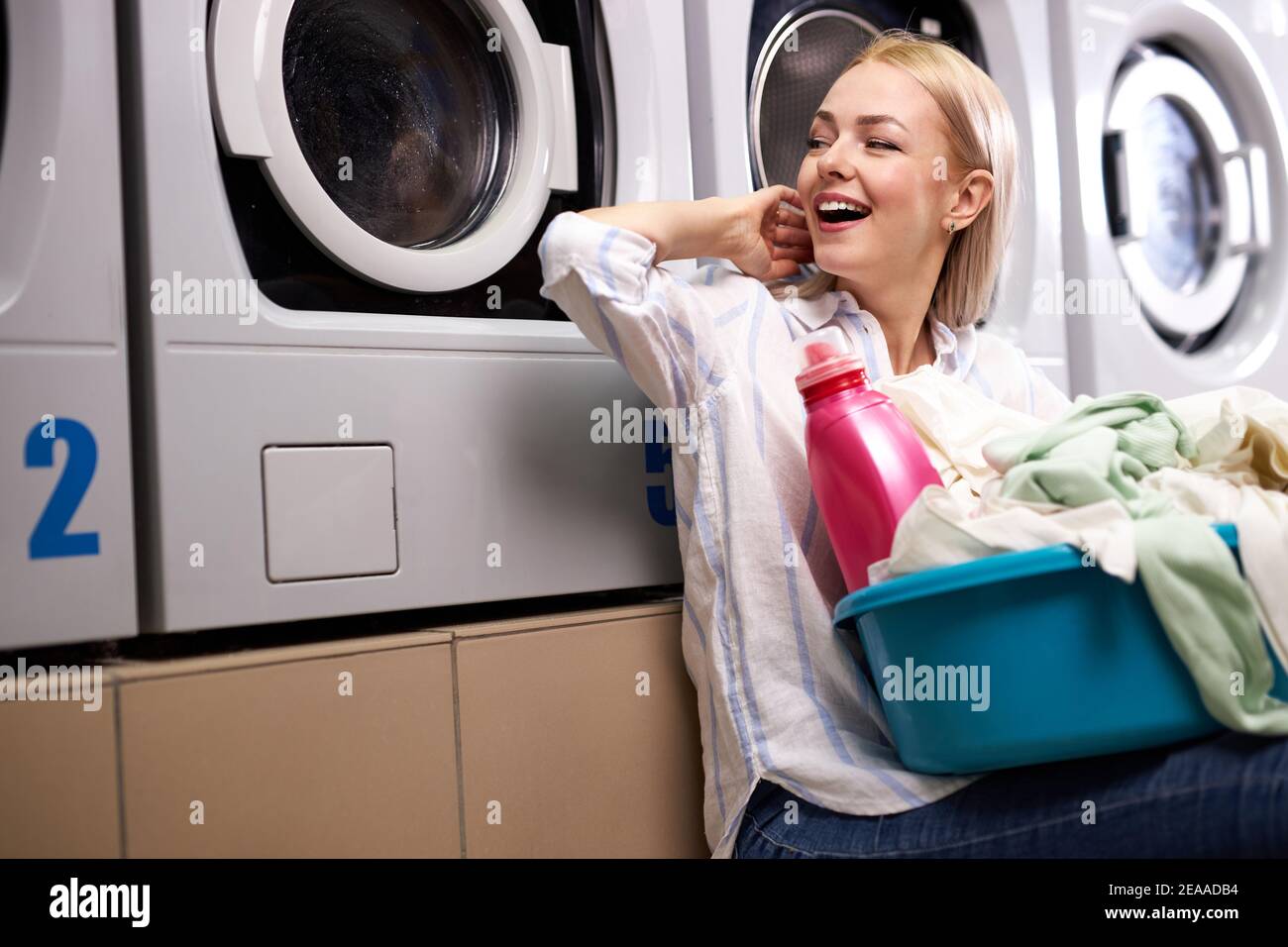 donna allegra si siede felicemente in attesa della fine della lavanderia, del sorriso, del bacino di presa con i vestiti sporchi e la bottiglia di detersivo rosa Foto Stock donna allegra si siede felicemente in attesa della fine della lavanderia, del sorriso, del bacino di presa con i vestiti sporchi e la bottiglia di detersivo rosa Foto Stock