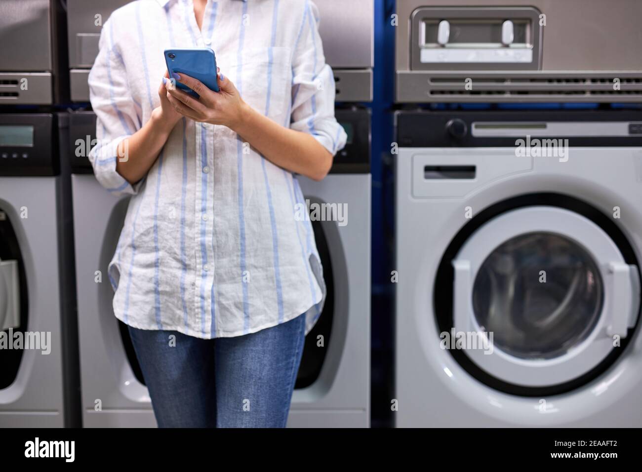 Facile processo di lavanderia. Foto ritagliata di giovane donna in piedi vicino alla lavatrice e guardando l'app per smartphone, in attesa di fine lavaggio Foto Stock