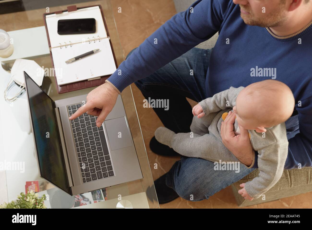 Il giovane padre lavora da casa mentre tiene il suo bambino sul suo grembo mentre entrambi guardano lo schermo. Foto Stock