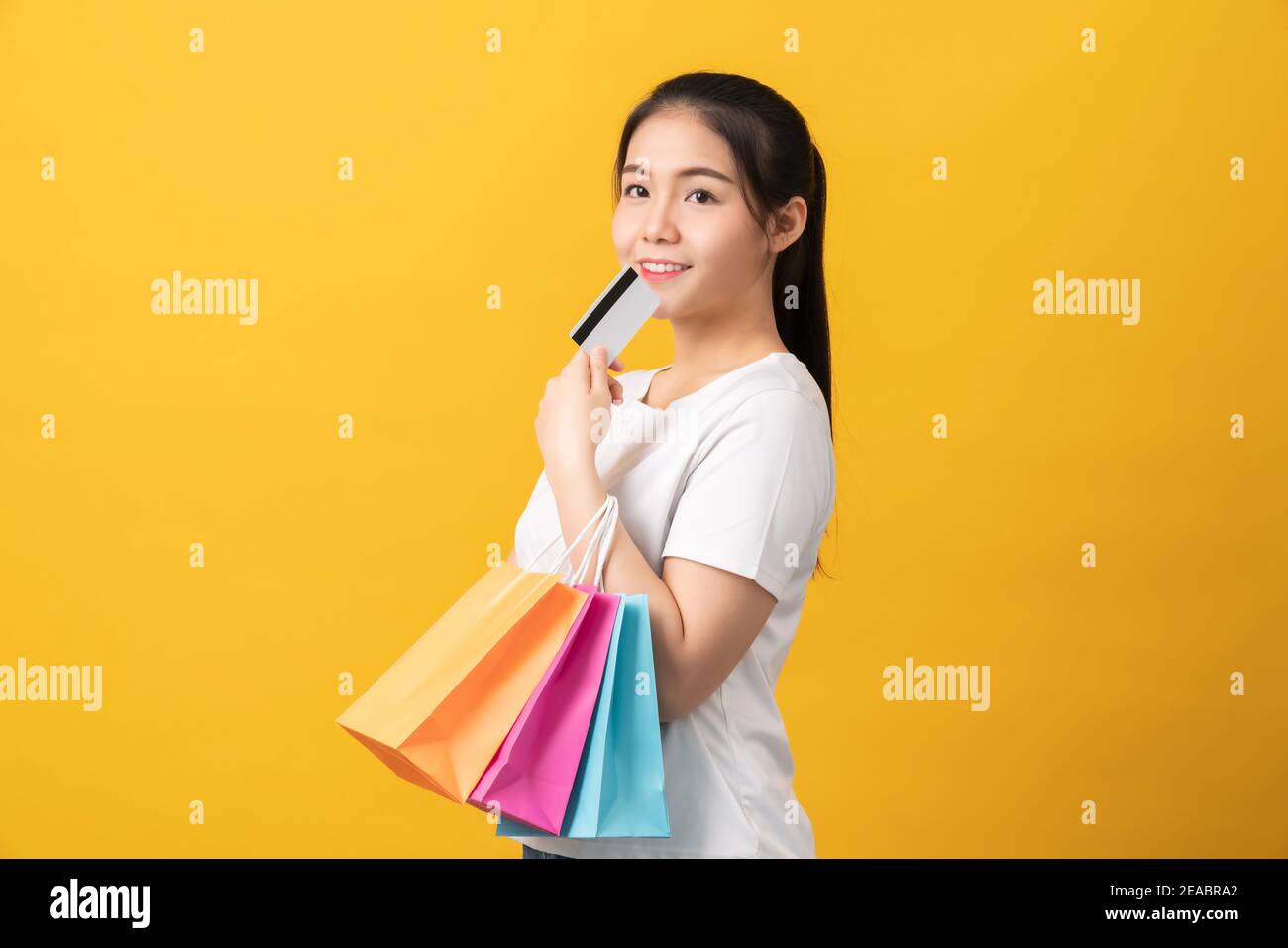 Allegra bella donna asiatica che tiene borse di shopping multicolore e carta di credito su sfondo arancione. Foto Stock